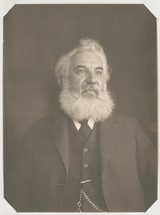 Sepia photograph of Alexander Graham Bell, a heavy-set man with white hair and a full white beard, in a dark three-piece suit with a watch chain, against a dark backdrop.