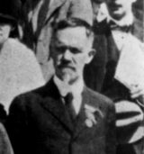Black-and-white head-and-shoulders detail of Charles B. Davenport, a man with short graying hair, a mustache and a short beard, in a dark suit with a badge on his lapel, standing among other people (detail of a group photograph captioned "Professor Moerns, Professor Shull, Dr. Davenport at the American Museum of Natural History").