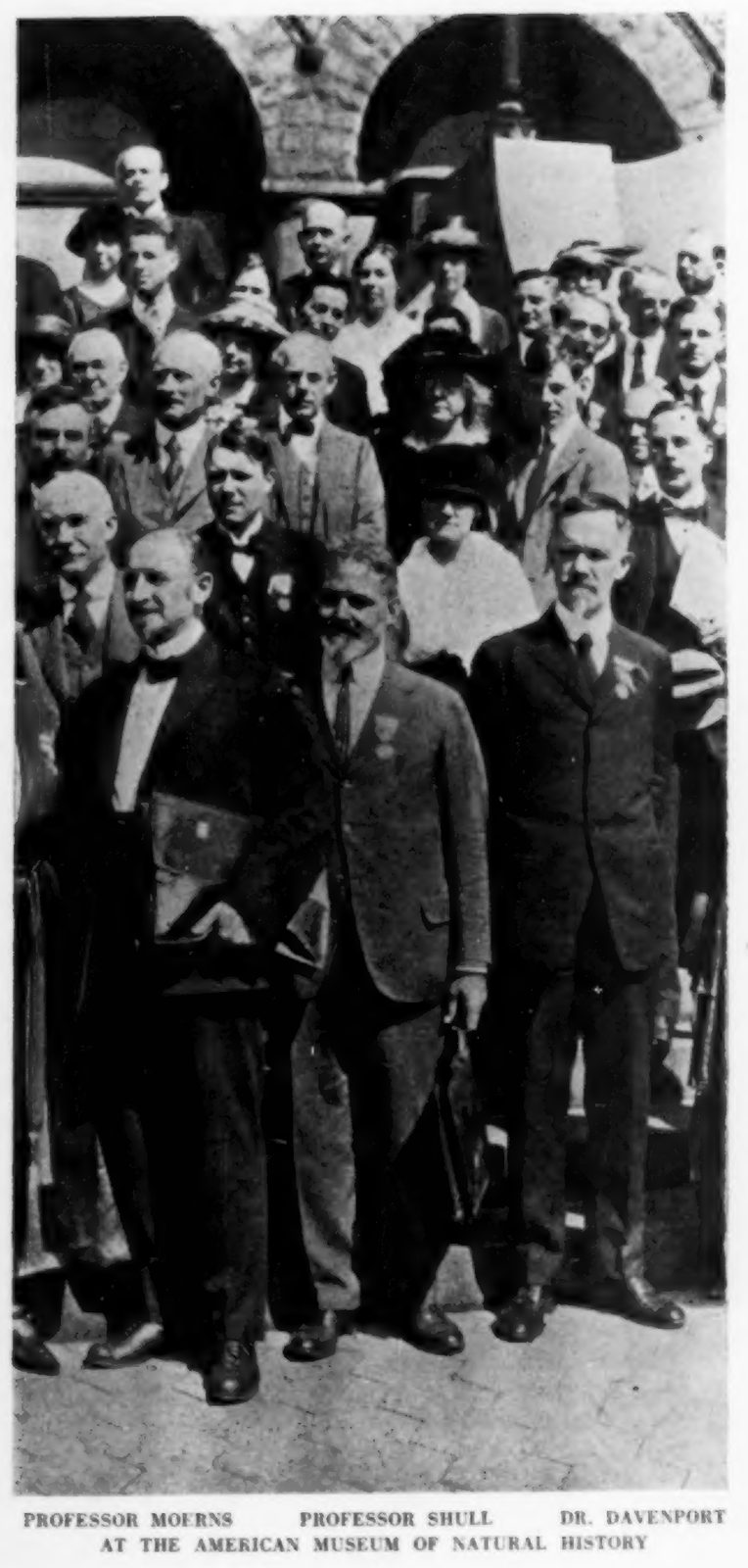 Black-and-white group photograph: three men in suits stand in front of a crowd of men and women on the steps of a stone building with arched doorways, captioned "Professor Moerns, Professor Shull, Dr. Davenport at the American Museum of Natural History," printed in The Scientific Monthly in November 1921 in its report on the Second International Congress of Eugenics, New York.